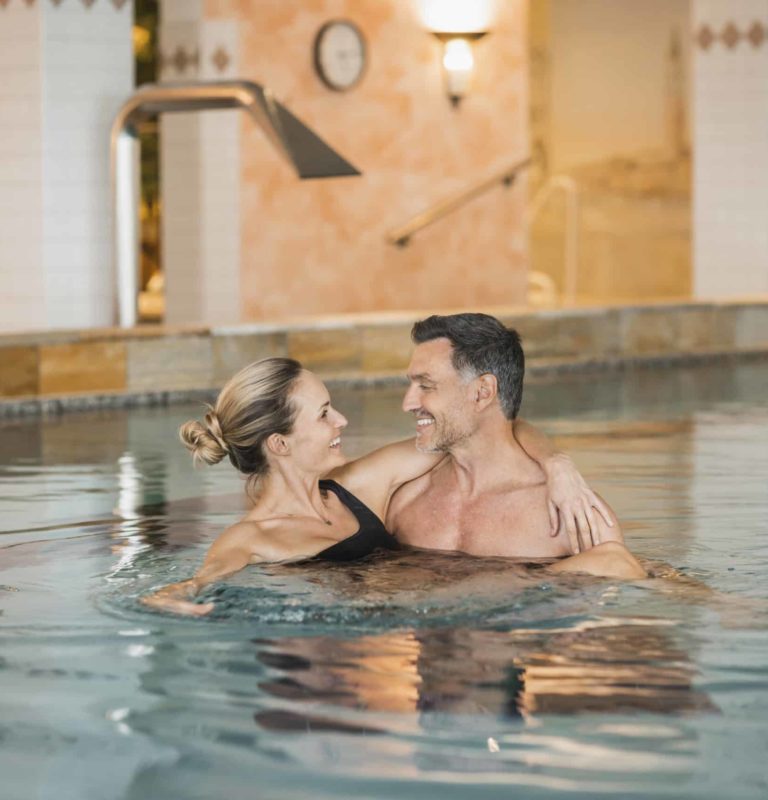 Frau liegt in den Armen ihres Mannes im indoor Pool. Sie lächeln sich an.